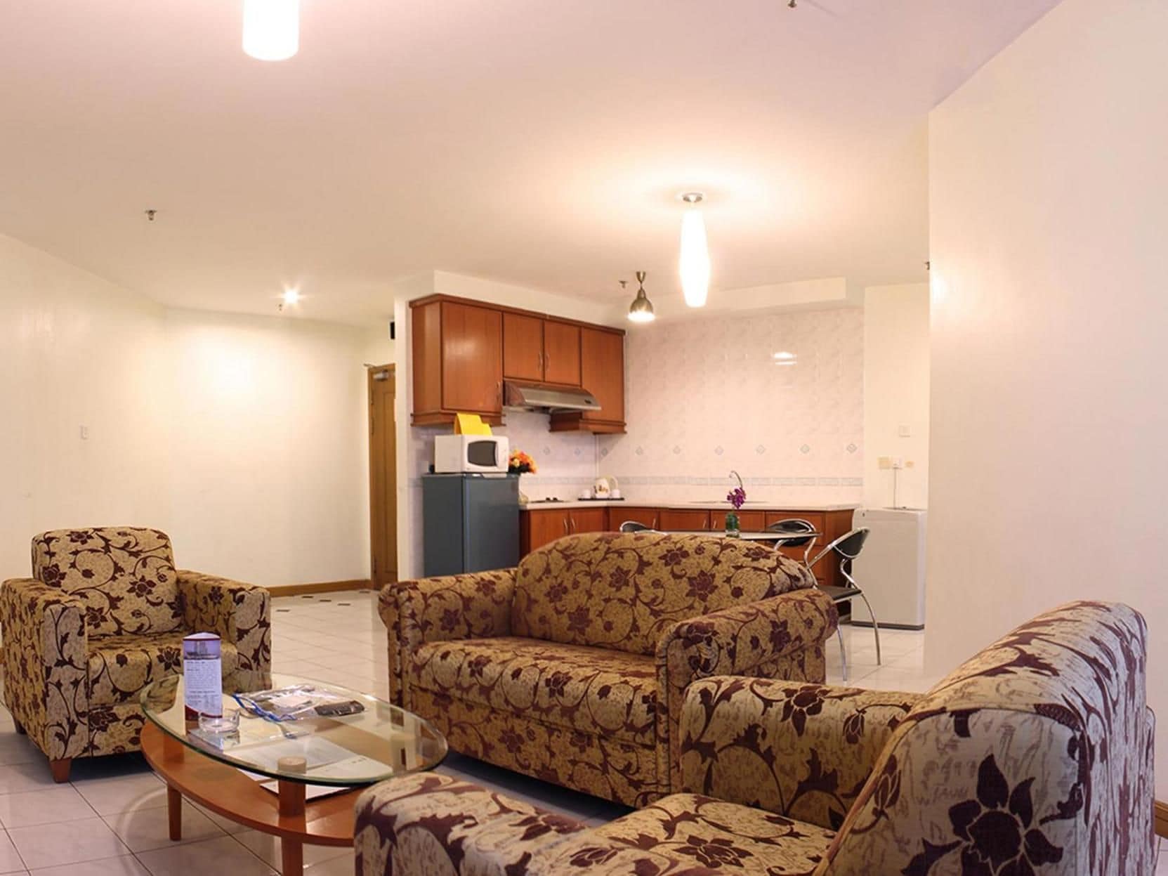 A living room with a sofa, armchairs, and a glass coffee table. There is a kitchen in the background with cabinets and a microwave.