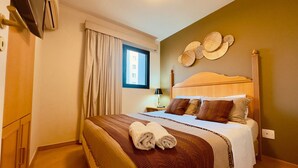 Habitación de hotel con cama, cortinas, ventana, televisor montado en la pared y mesita de noche con lámpara.