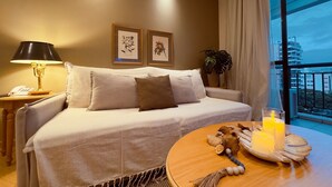Habitación de hotel con sofá, mesita de noche con lámpara, teléfono y ventana con vistas a edificios.