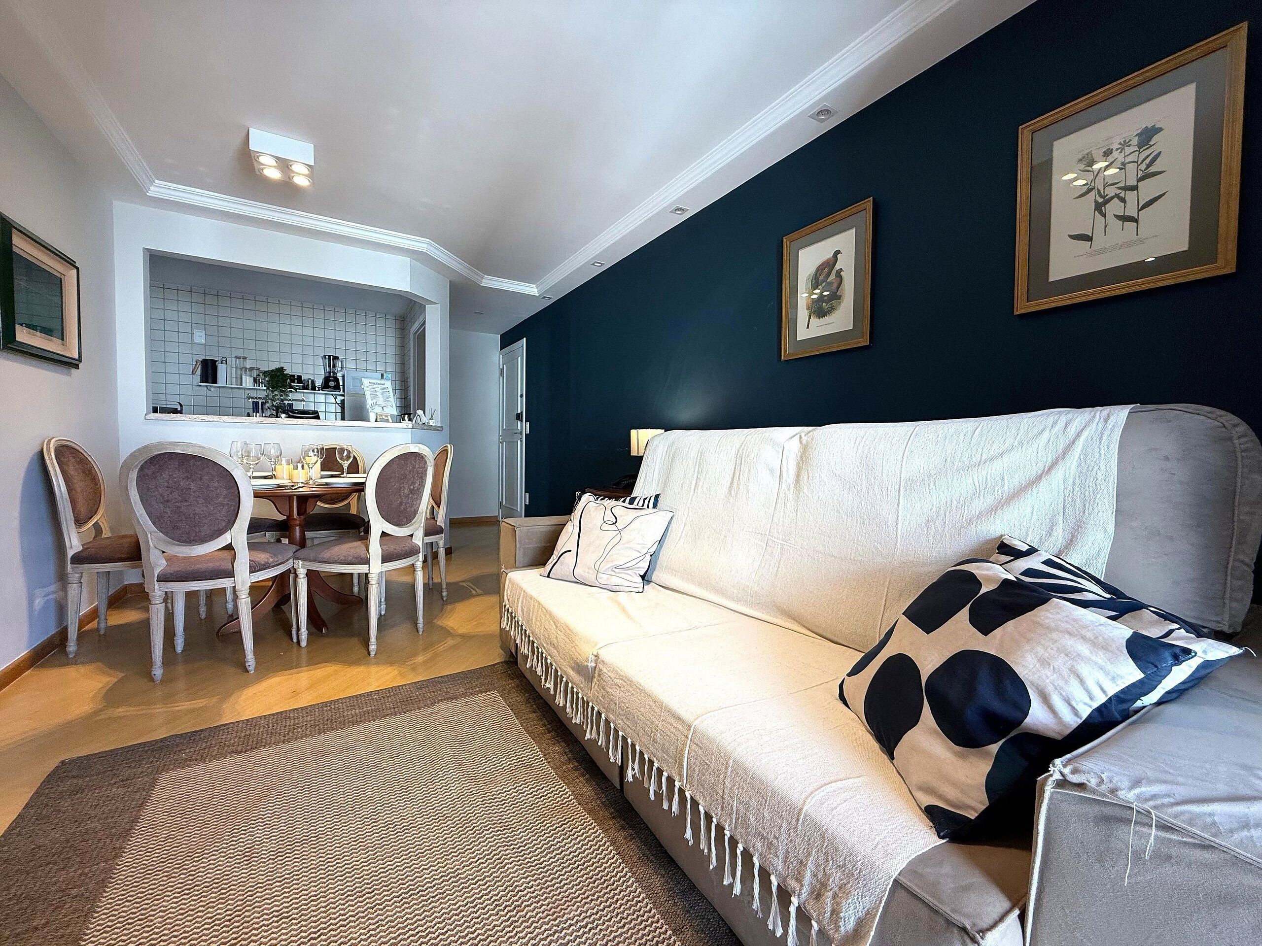 A living room with a white sofa, a dining table with chairs, and a kitchen area visible through an open door.