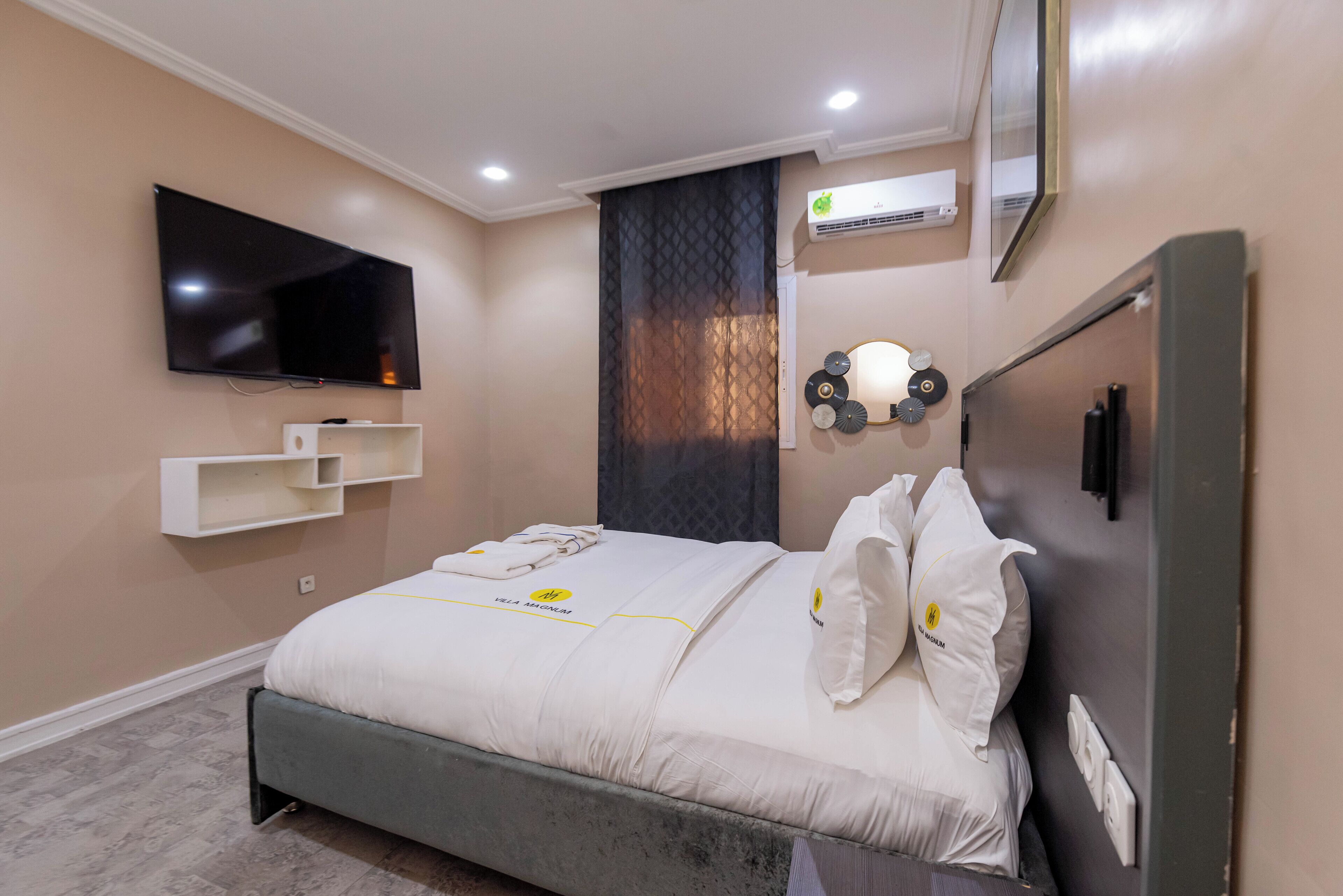 A modern bedroom with a bed, a mounted TV, a shelf, and an air conditioning unit.