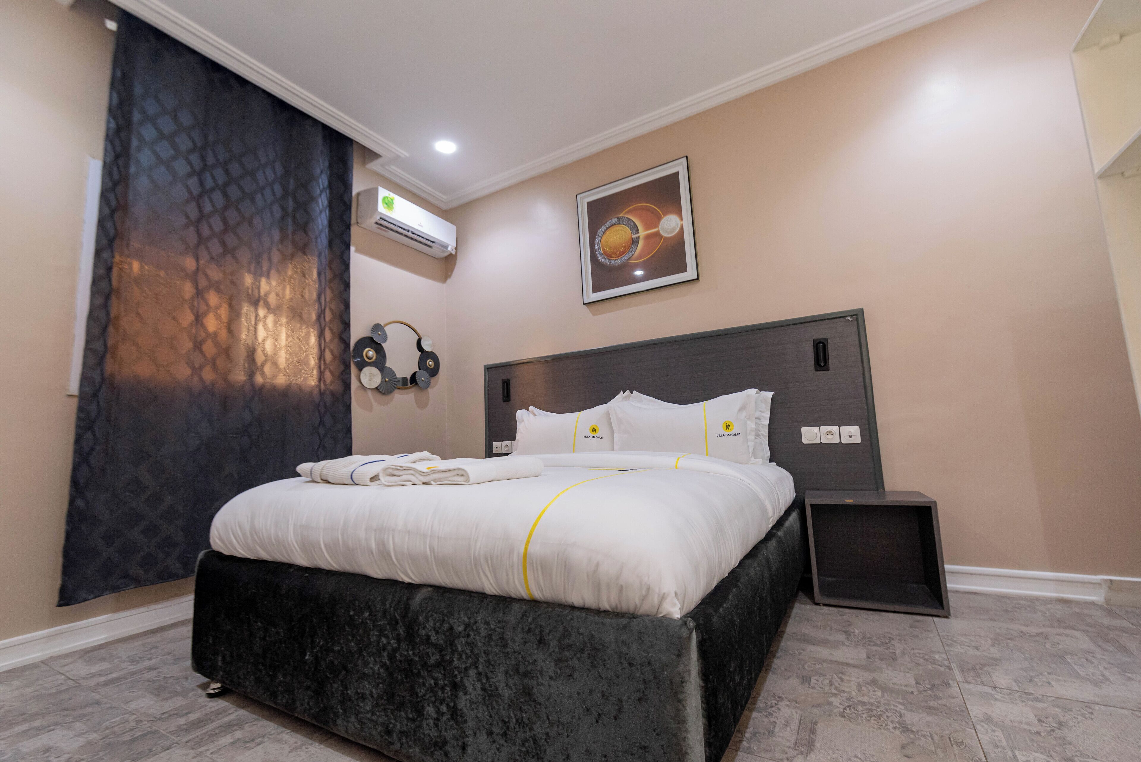 A neatly made bed with white linens and a black headboard, a wall-mounted air conditioner, and a framed picture of a planet.