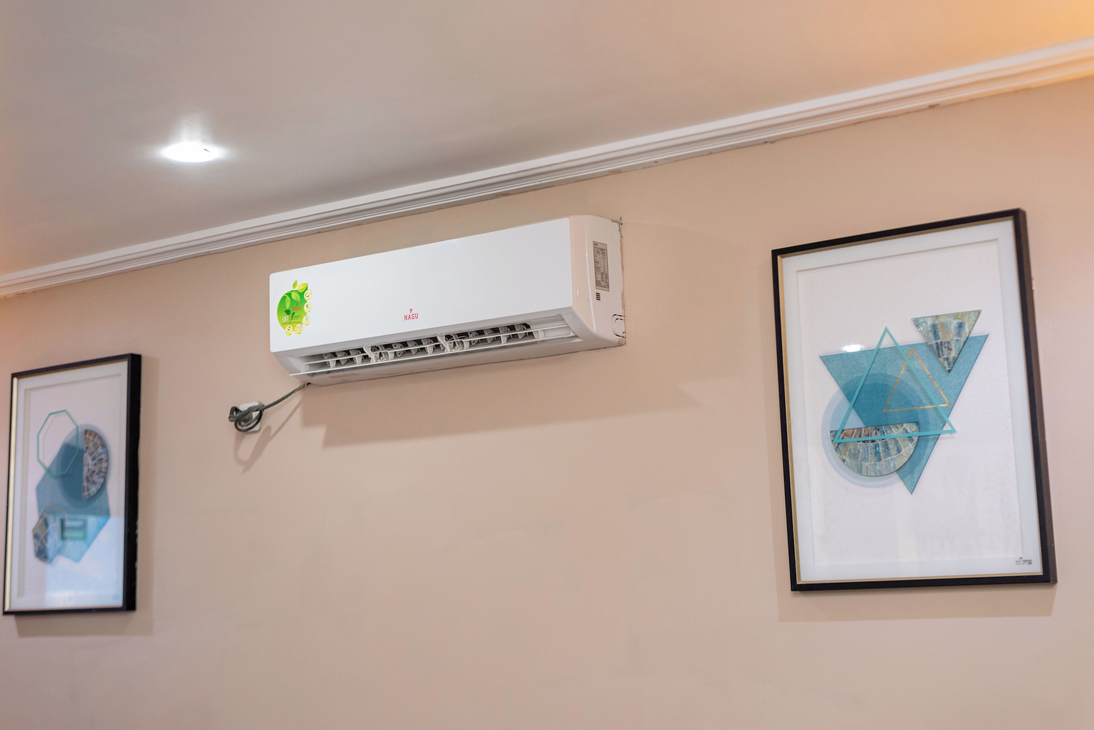 Un climatiseur mural avec un logo vert, deux œuvres d’art encadrées et un spot encastré au plafond.