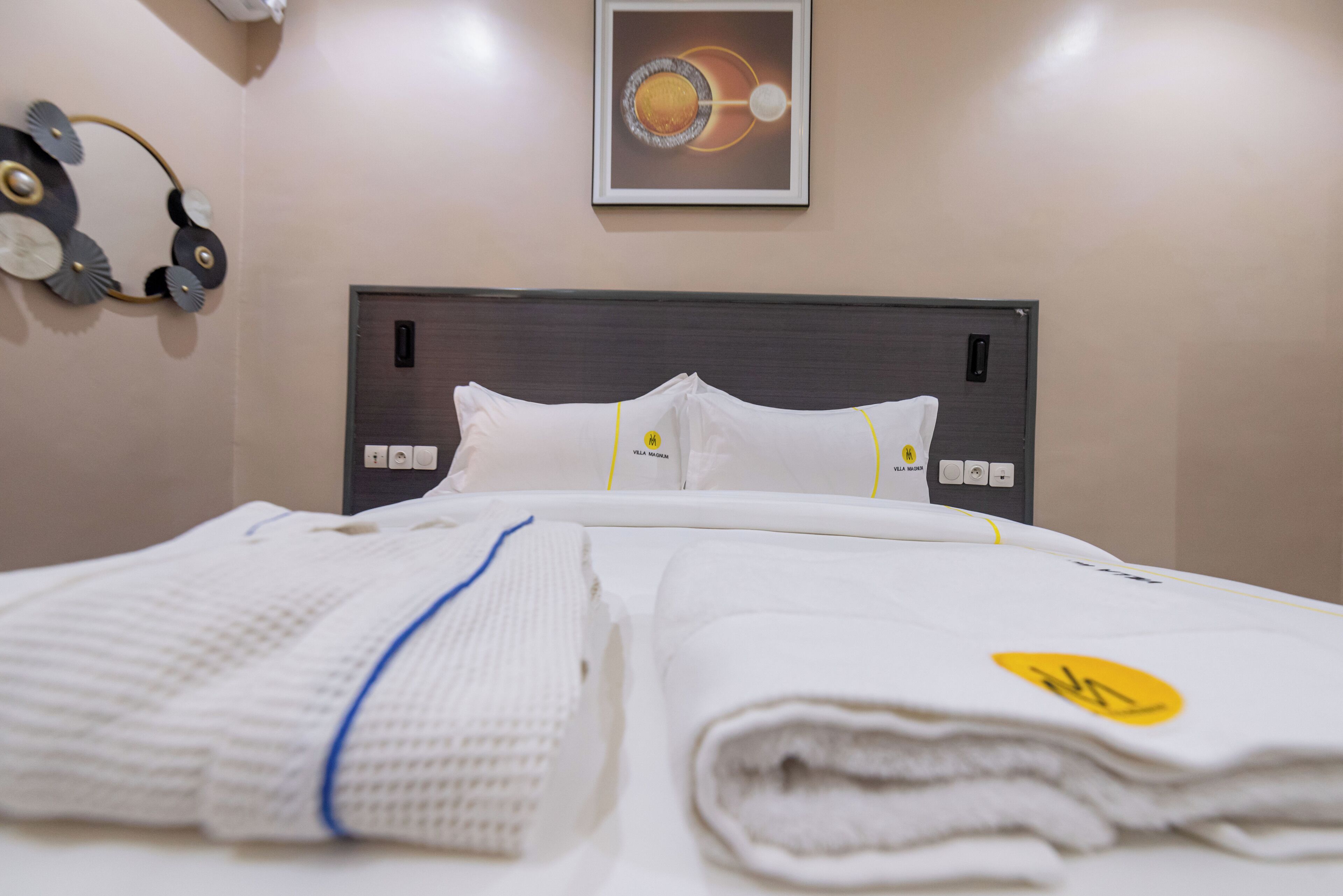 A neatly made bed with white linens and a folded towel, a wall clock, and a framed picture above the bed.