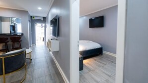 A modern hotel room with a bed, a TV mounted on the wall, a small desk with a chair, and a kitchen area visible through an open door. - Villa Magnum (Douala)