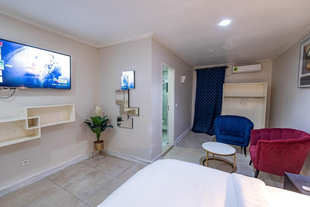 A hotel room with a bed, a TV mounted on the wall, a small table, two chairs, a potted plant, and a wardrobe.