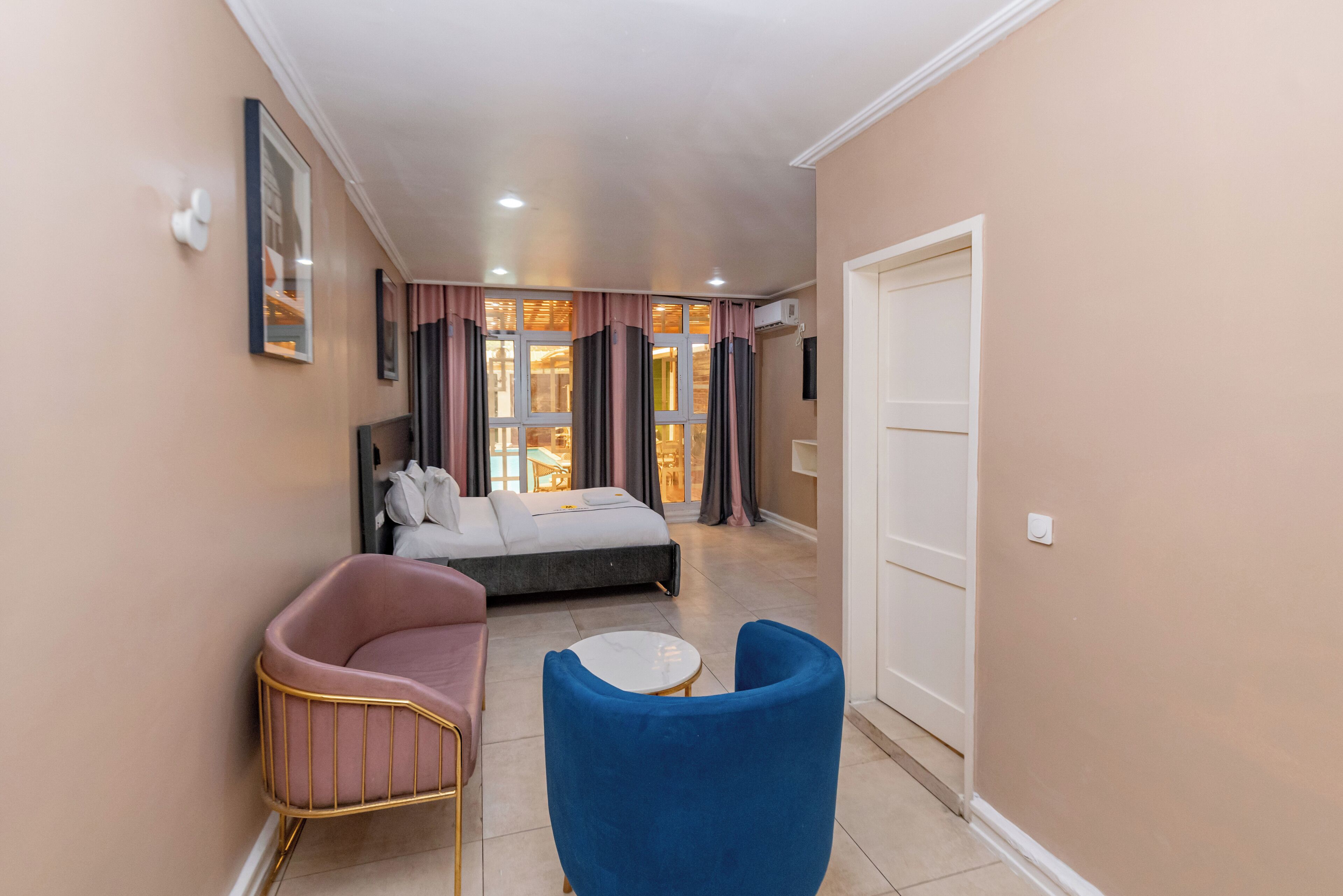 Une chambre d’hôtel avec un lit, un fauteuil rose, un fauteuil bleu et une petite table ronde.