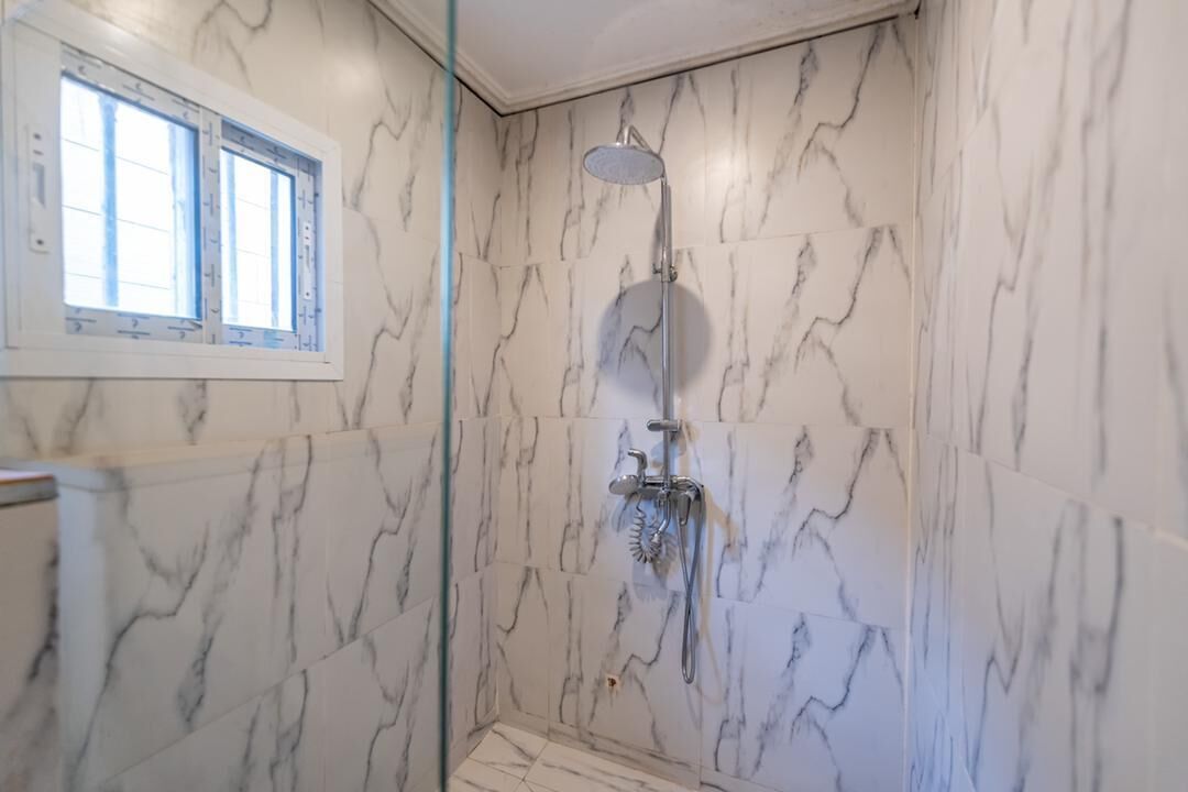 A bathroom with marble walls, a shower with a handheld showerhead, and a window with blinds.