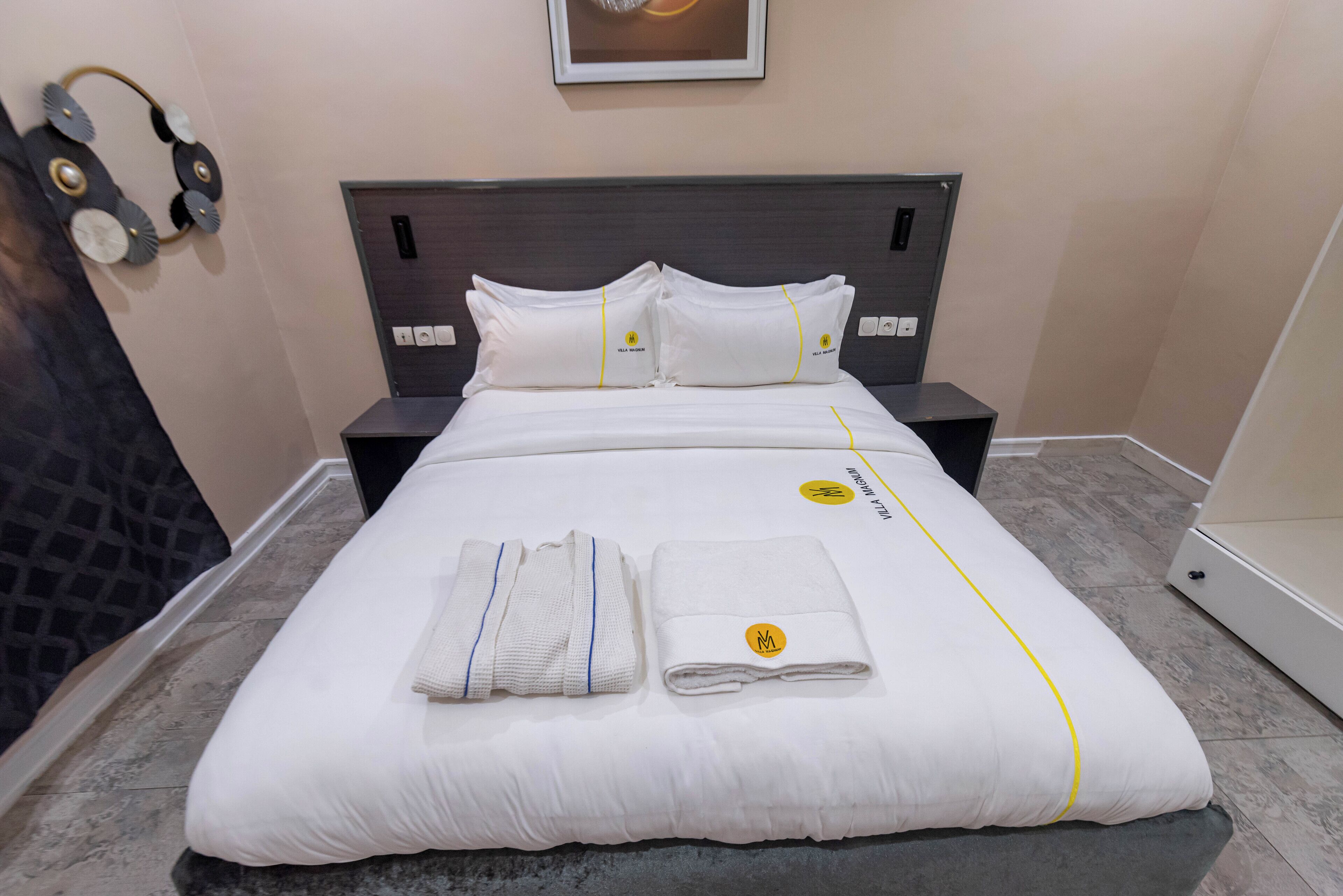 A neatly made bed with white bedding and pillows, a folded towel, and a pair of headphones on the wall.
