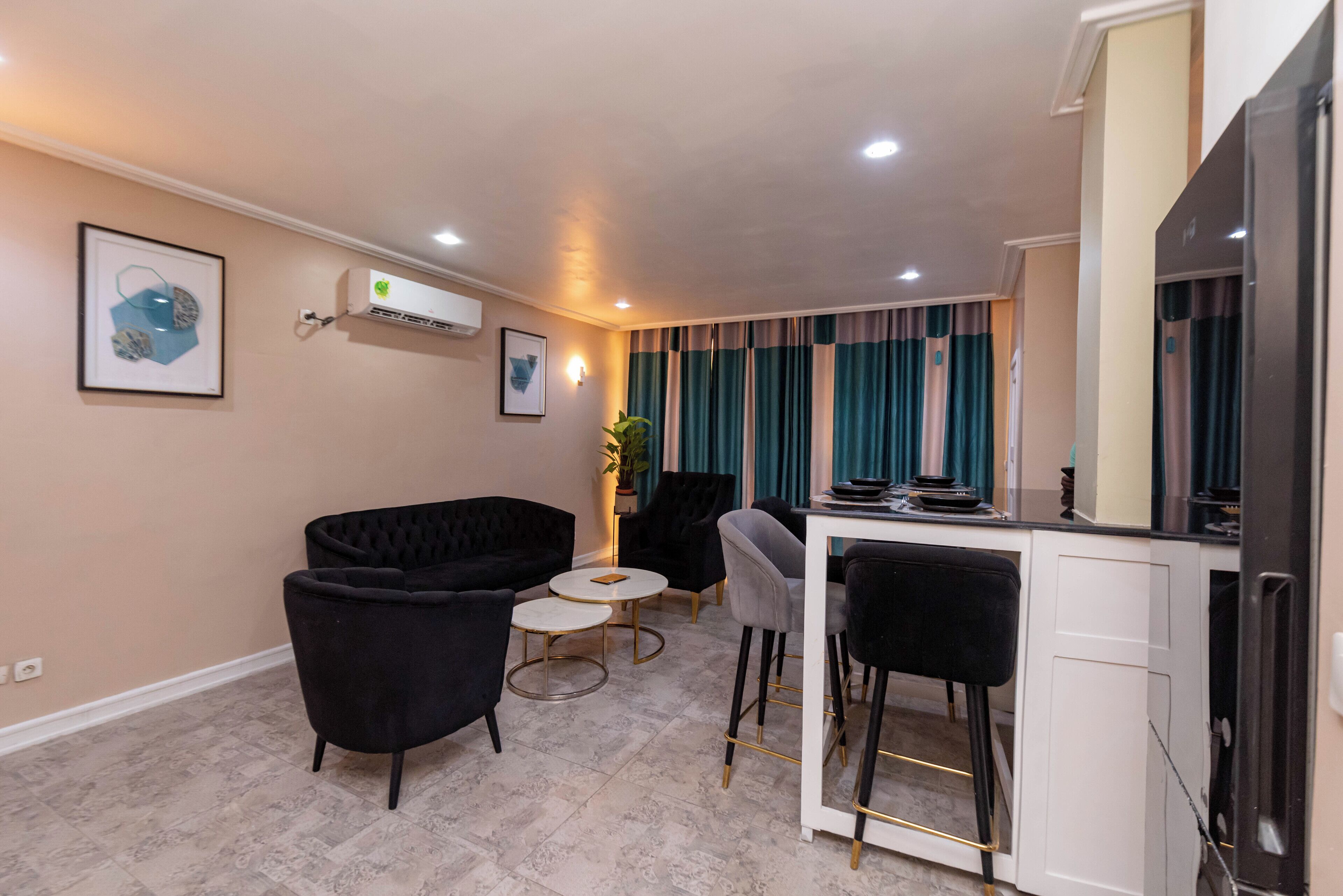 Un salon moderne avec un canapé noir, une table basse ronde et un coin bar comprenant un meuble blanc et un téléviseur.