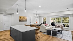 Eine moderne Küche mit Kochinsel, Essbereich mit Tisch und Stühlen, ein Wohnzimmer mit Sofa und Couchtisch sowie große Fenster mit Blick auf Grünflächen.