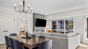 A modern living room with a grey sofa, a wooden dining table with chairs, a flat-screen TV mounted on the wall, and a large window offering a view of the outdoors. - 3 Bed/2 Bath Sparrowhawk Lodge-South Facing (Dead Man's Flats)