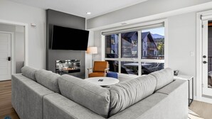 A modern living room with a grey sectional sofa, a round coffee table, a flat-screen TV mounted on the wall, and a large window offering a view of a mountain and a building. - 3 Bed/2 Bath Sparrowhawk Lodge-South Facing (Dead Man's Flats)
