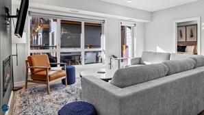 A modern living room with a grey sofa, a brown leather chair, and a blue ottoman. There is a flat-screen TV mounted on the wall, a small side table, and a large window providing a view of the outside. - 3 Bed/2 Bath Sparrowhawk Lodge-South Facing (Dead Man's Flats)