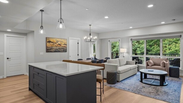 A modern kitchen with a large island, dining area with a sofa and chairs, and a living room with a coffee table and a view of greenery through large windows.
