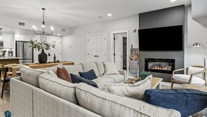 A modern living room with a large sectional sofa, a flat-screen TV mounted on the wall, and a fireplace.