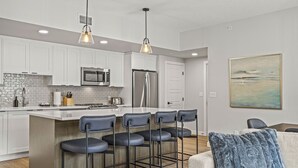 Uma cozinha moderna com armários brancos, uma ilha central com banquetas, eletrodomésticos de aço inoxidável e iluminação pendente.