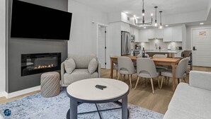 Un salon moderne avec une cheminée, un téléviseur à écran plat, un coin repas avec des chaises et une cuisine en arrière-plan.