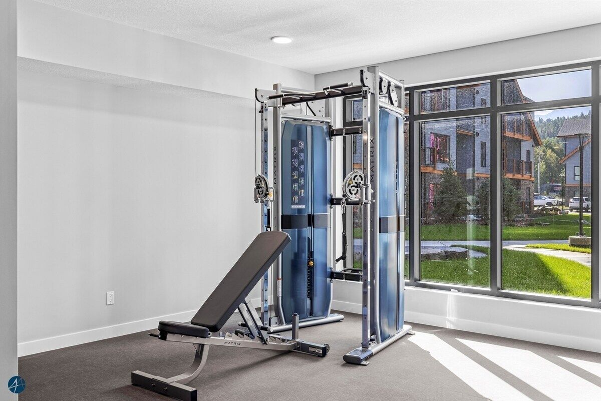 Une salle de sport équipée de matériel de musculation et d’un banc, de grandes fenêtres offrant une vue sur les bâtiments et la verdure.