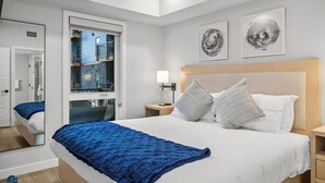 Une chambre avec un grand lit, une table de chevet, une fenêtre donnant sur un immeuble et des œuvres d’art accrochées au mur.