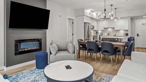 Un salon moderne doté d’un téléviseur à écran plat, d’une cheminée, d’un coin repas avec une table et des chaises, et d’une cuisine en arrière-plan.