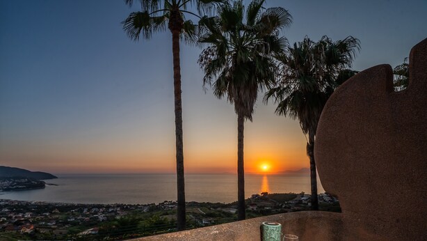 Ein Tisch mit Essen und einem Weinglas, mit Blick auf einen Sonnenuntergang über dem Meer mit Palmen.