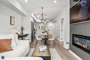 Una sala de estar moderna con sofá, mesa de centro y chimenea. Hay un comedor con mesa y sillas, y una cocina al fondo.