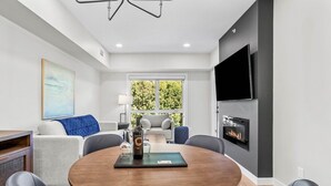 A modern living room with a round wooden table, comfortable seating, a fireplace, a large window, and a flat-screen TV mounted on the wall. - The Creekside - Top Floor Mountiain & Creek Views (Dead Man's Flats)