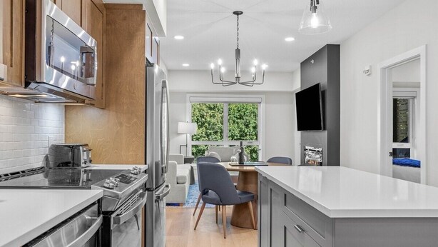 A modern kitchen with stainless steel appliances, a large island, and a dining area with a view of greenery. - The Creekside - Top Floor Mountiain & Creek Views (Dead Man's Flats)