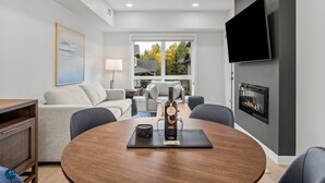 A modern living room with a round wooden table, a fireplace, a flat-screen TV, and a large window with a view of trees.