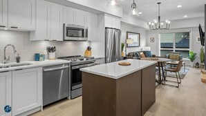 Une cuisine moderne avec des meubles blancs, des appareils en acier inoxydable, un îlot central et des luminaires suspendus.