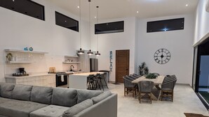 Uma cozinha moderna com uma ilha central, uma sala de jantar com mesa e cadeiras de madeira, e um sofá modular cinza.