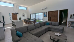 Uma sala de estar espaçosa com um amplo sofá em formato de L, uma área de jantar com mesa e cadeiras, e vista para o exterior através de grandes janelas.
