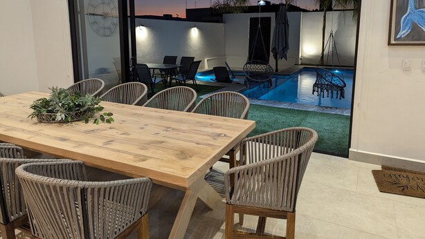 Área de refeições interna com mesa de madeira e cadeiras de vime, vista para uma piscina e porta que dá para o exterior.