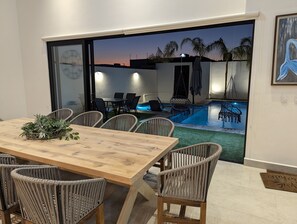 Un'area pranzo interna con tavolo in legno e sedie in vimini, vista su una piscina e una porta che dà all'esterno.