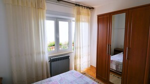 Une chambre avec un lit, un radiateur, une fenêtre avec des rideaux, une armoire et une vue sur le balcon extérieur.