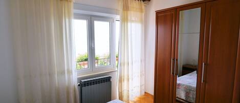 A bedroom with a bed, a radiator, a window with curtains, a wardrobe, and a view of the outside balcony.