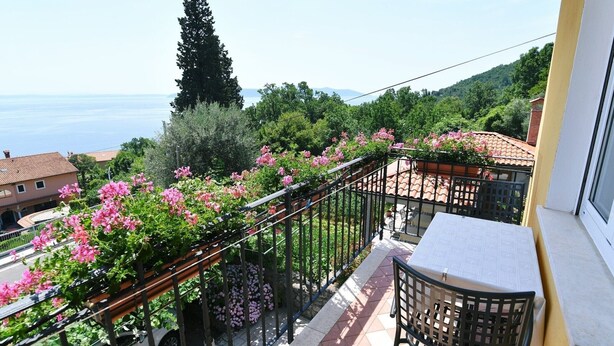 A balcony with a table and chair, overlooking a garden with pink flowers and a view of the sea.