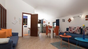 A modern living room with a blue sofa, a dining area with orange chairs, and a kitchen in the background.