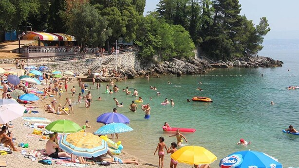 A crowded beach with people sunbathing and swimming, surrounded by trees and a rocky shoreline.