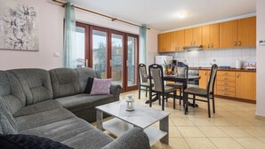 Ein Wohnzimmer mit grauem Sofa, einem Couchtisch und einem Essbereich mit Tisch und Stühlen. Es gibt eine Küche mit Holzschränken und einem Fenster mit Blick auf einen Balkon.