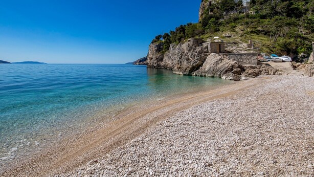 Une plage de galets bordée d’une eau claire, une falaise rocheuse et un bâtiment perché au sommet.