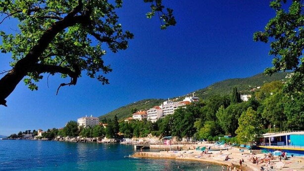 Eine Küstenlandschaft mit Strand, klarem blauen Wasser und Gebäuden an einem Hügelhang.