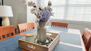 Une table à manger ornée d’un vase contenant des plantes de coton et des fleurs violettes, un plateau en bois avec un bocal et des perles, et une lampe en arrière-plan.