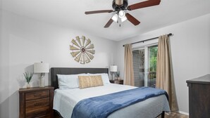 Une chambre à coucher comprenant un lit, des tables de chevet, un ventilateur de plafond, un élément décoratif mural et une vue à travers une porte-fenêtre coulissante.