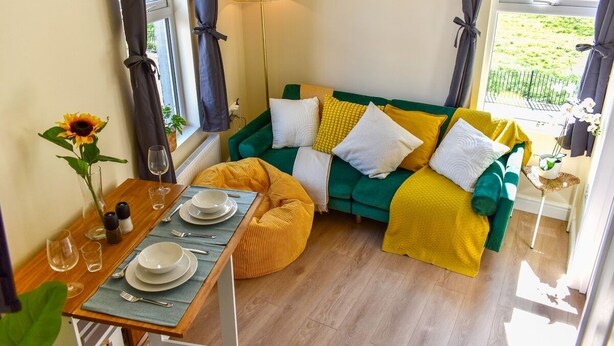Ein gemütliches Wohnzimmer mit einem grünen Sofa, gelben Kissen, einem Esstisch aus Holz und einer Stehlampe.