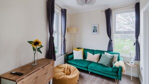 Ein Zimmer mit einem grünen Sofa, einer Holzkommode, einem gelben Hocker, einer Vase mit einem Sonnenblume und einem Fenster mit Vorhängen.