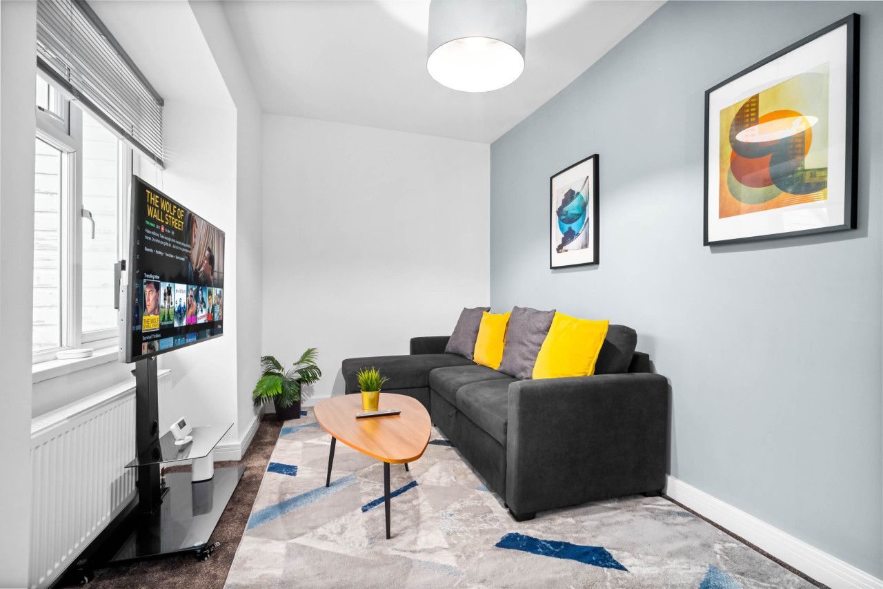 Un salon moderne avec un canapé gris, des coussins jaunes, une table basse et un téléviseur fixé au mur qui affiche une affiche de film.
