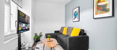 Un salon moderne avec un canapé gris, des coussins jaunes, une table basse et un téléviseur fixé au mur qui affiche une affiche de film.