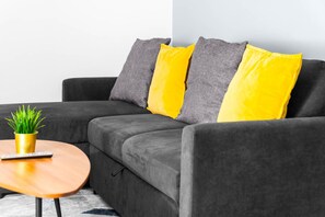 A grey sofa with yellow cushions, a small wooden table with a plant and a remote control, and a grey rug.